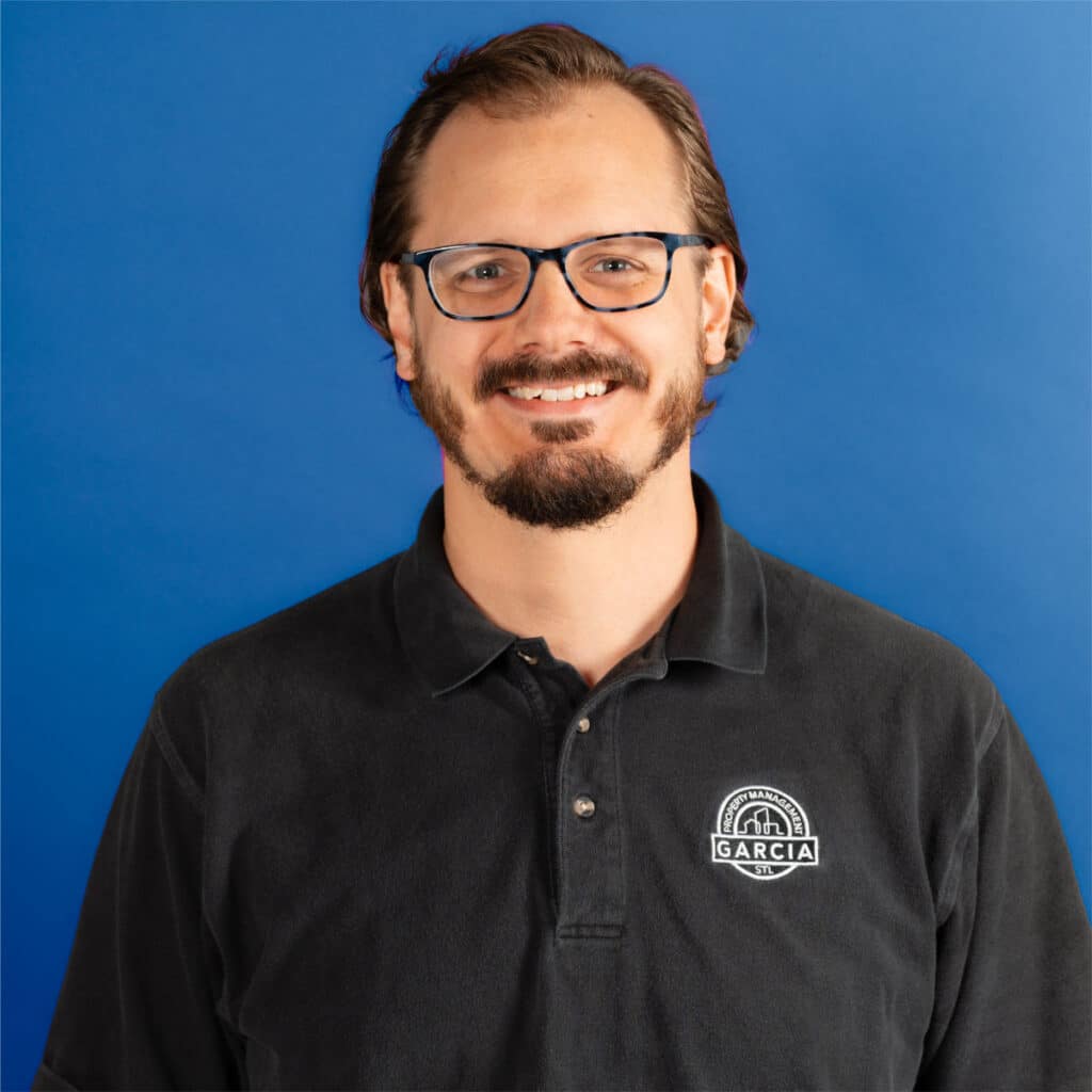 Smiling man with short brown hair, beard, and glasses wearing a black polo shirt with the “Garcia Property Management” logo, standing in front of a blue background.