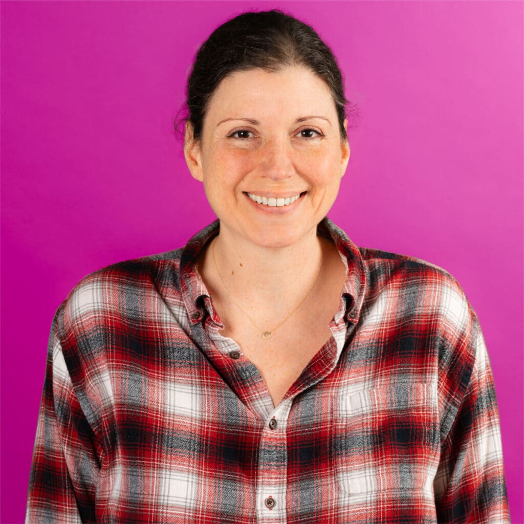 Smiling woman with dark hair pulled back, wearing a red, white, and black plaid shirt, standing in front of a bright pink background.