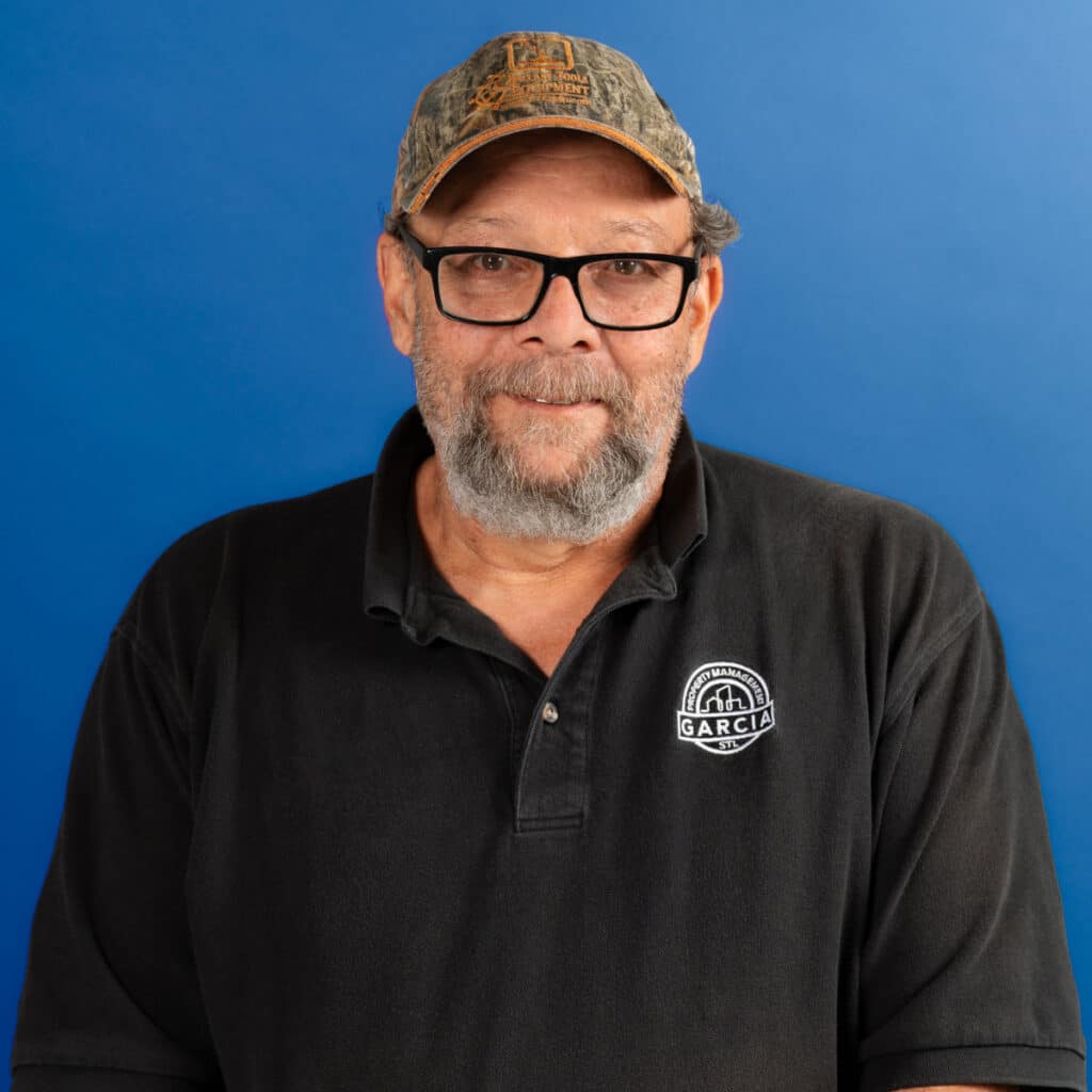 Smiling older man with glasses and a gray beard, wearing a camouflage baseball cap and a black polo shirt with a “Garcia Property Management STL” logo. He stands in front of a solid blue background.