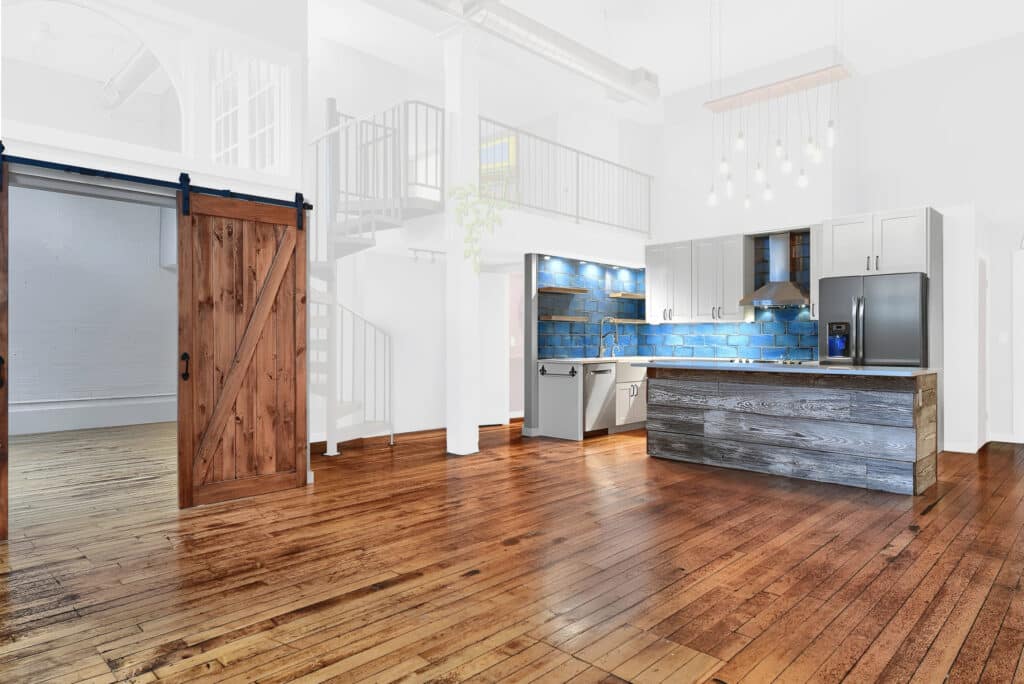 Open-concept loft with a modern-rustic kitchen and hardwood floors. The kitchen features a reclaimed wood island, blue tile backsplash, stainless steel appliances, white upper cabinets, and open wooden shelves. A sliding barn door made of natural wood reveals an adjacent room with exposed brick walls and large windows. The space is bright and airy with white walls, high ceilings, and a spiral staircase leading to a mezzanine level. Hanging pendant lights add a contemporary touch.